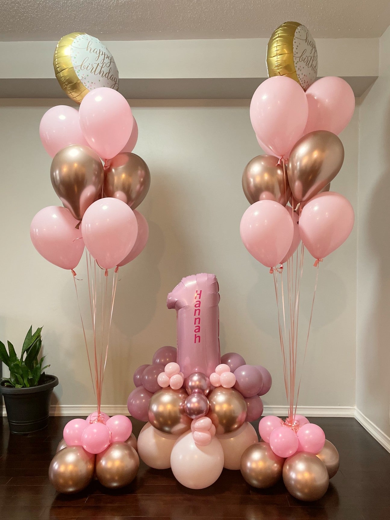 Two pink and gold balloon bunches with a bouquet in the middle with the number one in it.